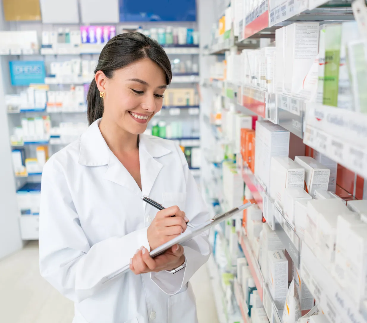 about-1 Smiling pharmacist in white coat writing on a clipboard in a pharmacy aisle with shelves of medicine.