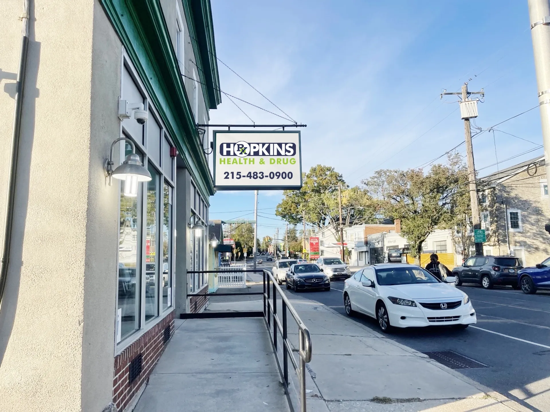 about-2 Street view of Hopkins Health & Drug pharmacy sign, with cars parked and driving along a sunny road.