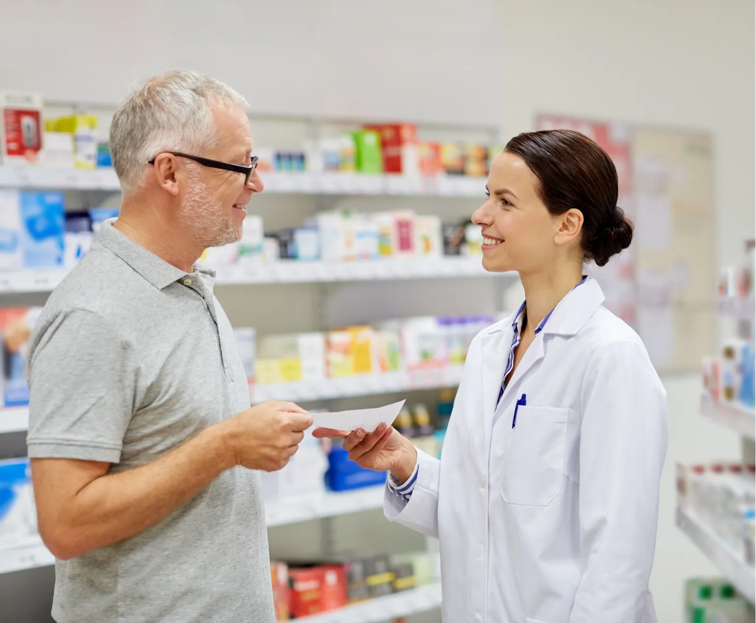A pharmacist in a white coat smiles while helping a man holding a prescription in a pharmacy.