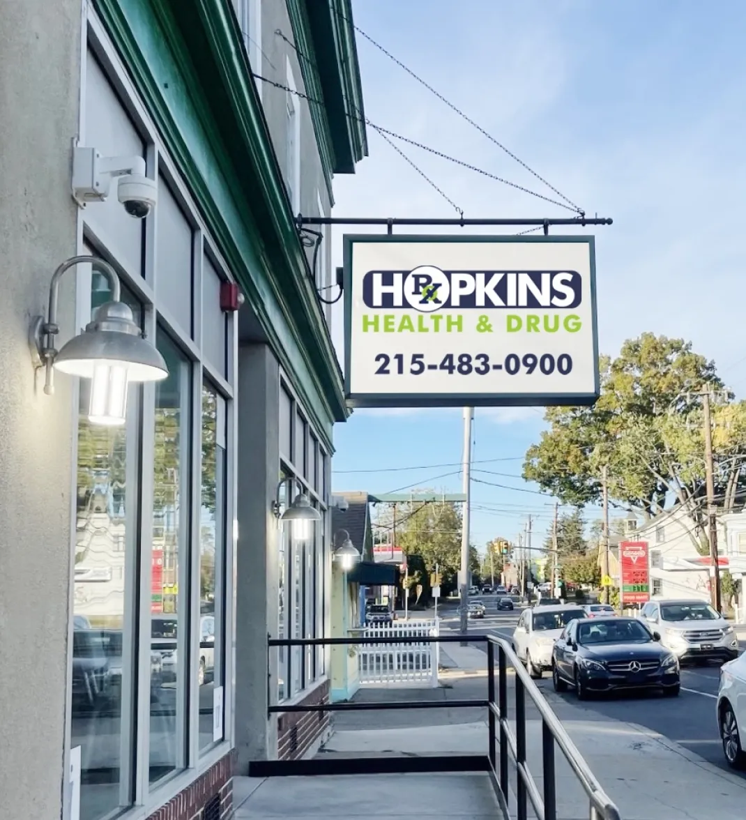 Street view of Hopkins Health & Drug pharmacy sign with phone number outside a building on a sunny day.