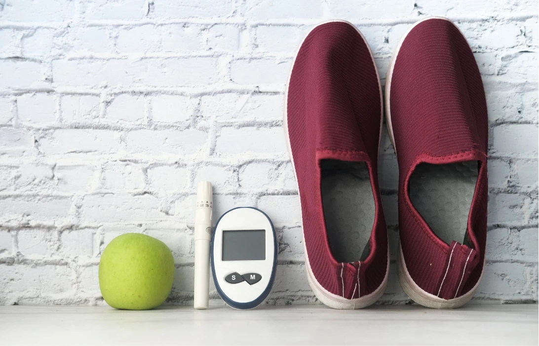 Green apple, glucose meter, test strip, and red sneakers lined up against a white brick wall.