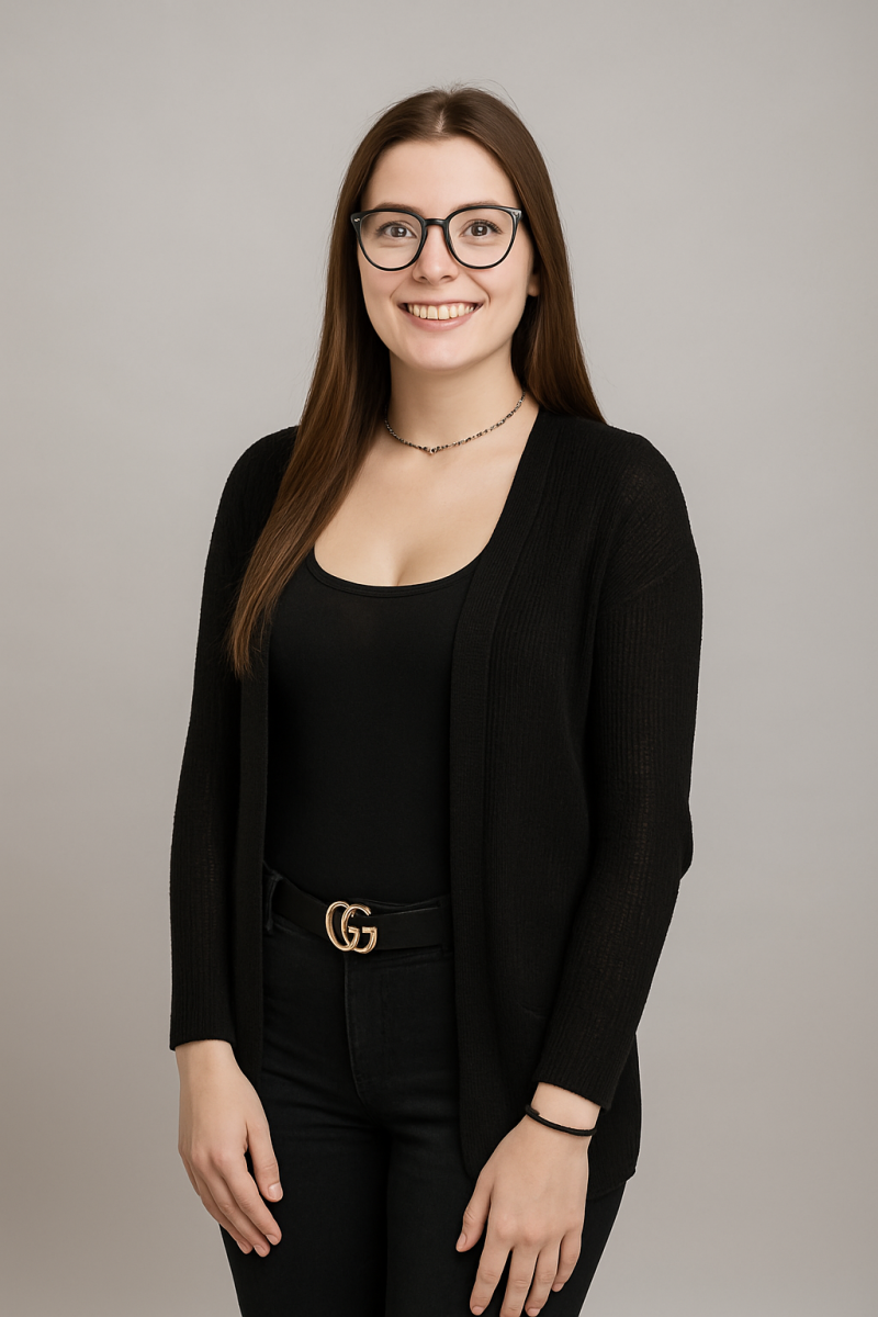Woman with long brown hair, wearing glasses and black outfit, smiling in front of a neutral gray background.