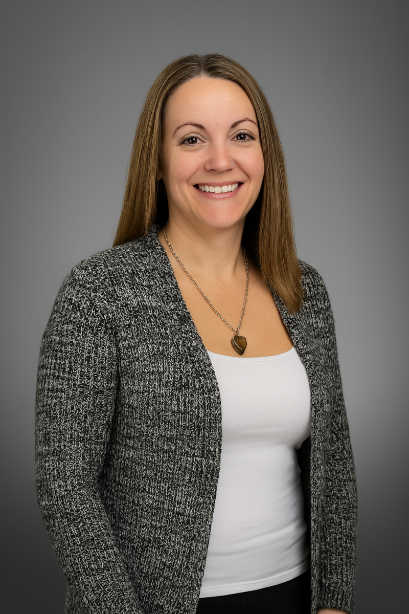 Smiling woman with straight brown hair, wearing a gray cardigan and white top, posing against a gray background.