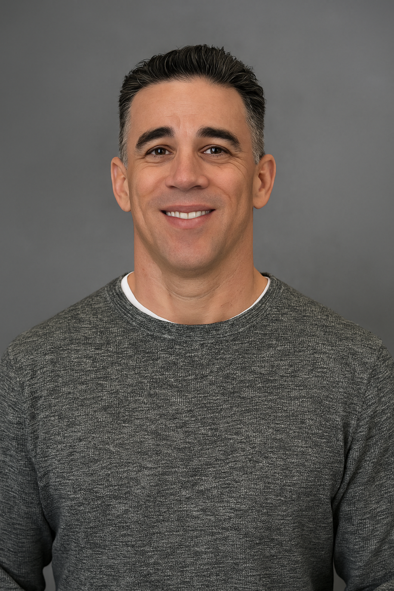 Man with short dark hair in a gray sweater, smiling in front of a plain gray background.