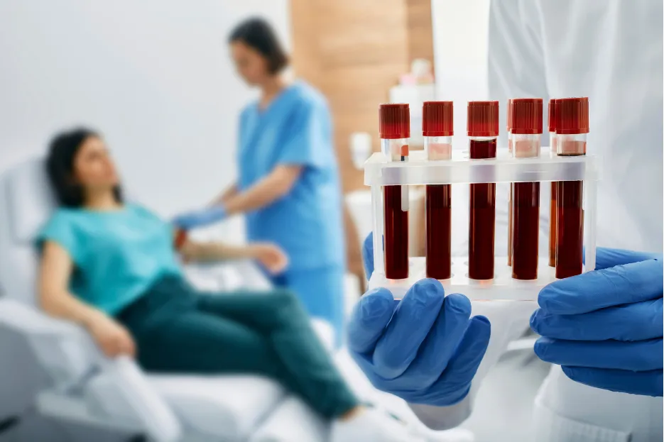 testing-1 A gloved hand holds blood samples, while a nurse attends to a patient in the background.