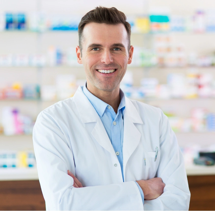 why-choose Smiling pharmacist in a white coat stands with arms crossed in a pharmacy.