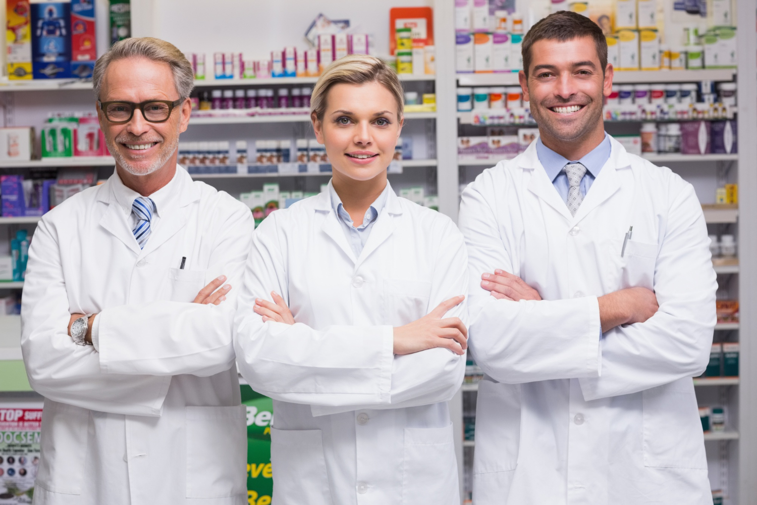 102282 Three pharmacists in white coats standing with arms crossed, smiling in a pharmacy.