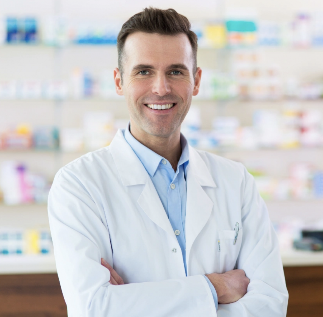 Smiling pharmacist in a white coat stands with arms crossed in a pharmacy with shelves of medicine behind him.
