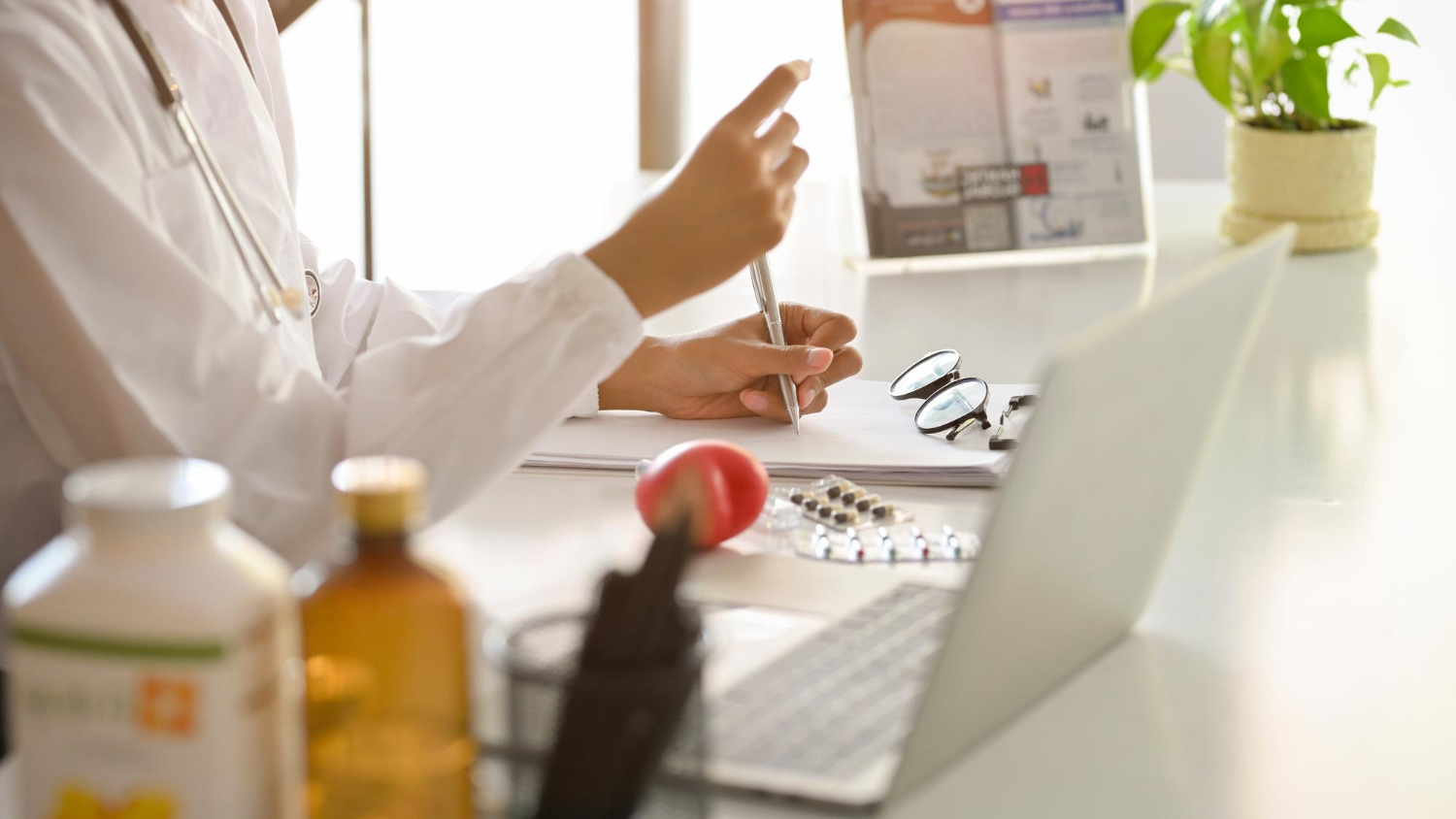 246775 A doctor writes notes at a desk with a laptop, glasses, medication, and a plant.