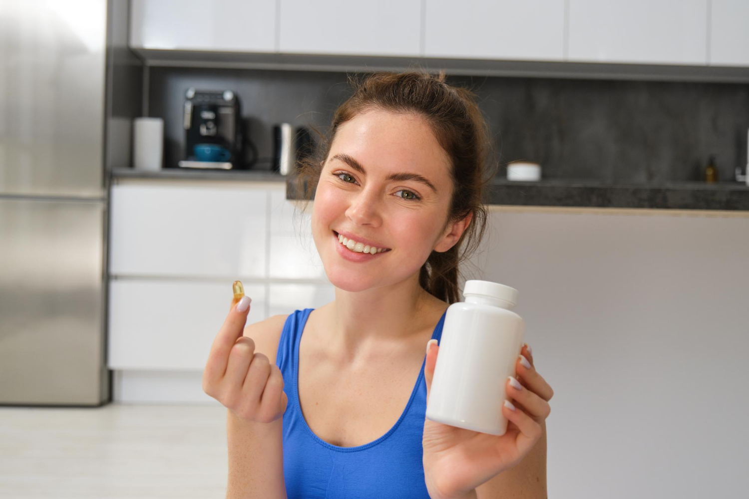 189563 Woman in blue top smiles, holding a white pill bottle and a single capsule in a modern kitchen.