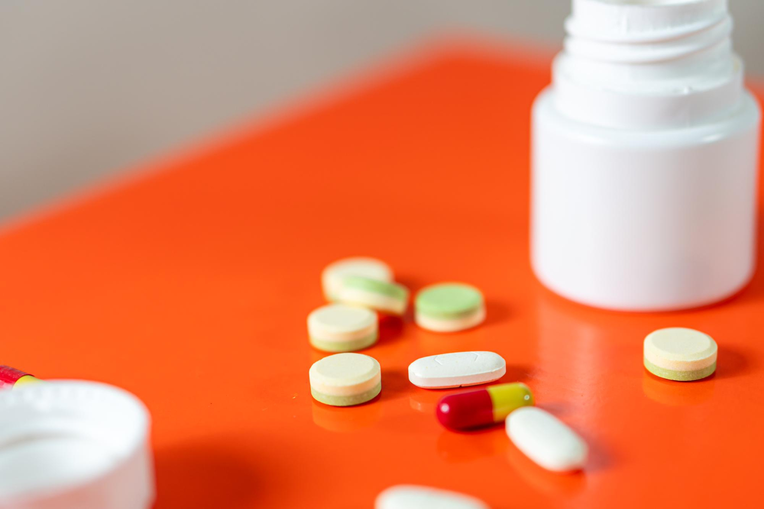 3686 Round yellow pills and a red-yellow capsule near an open white pill bottle on an orange surface.