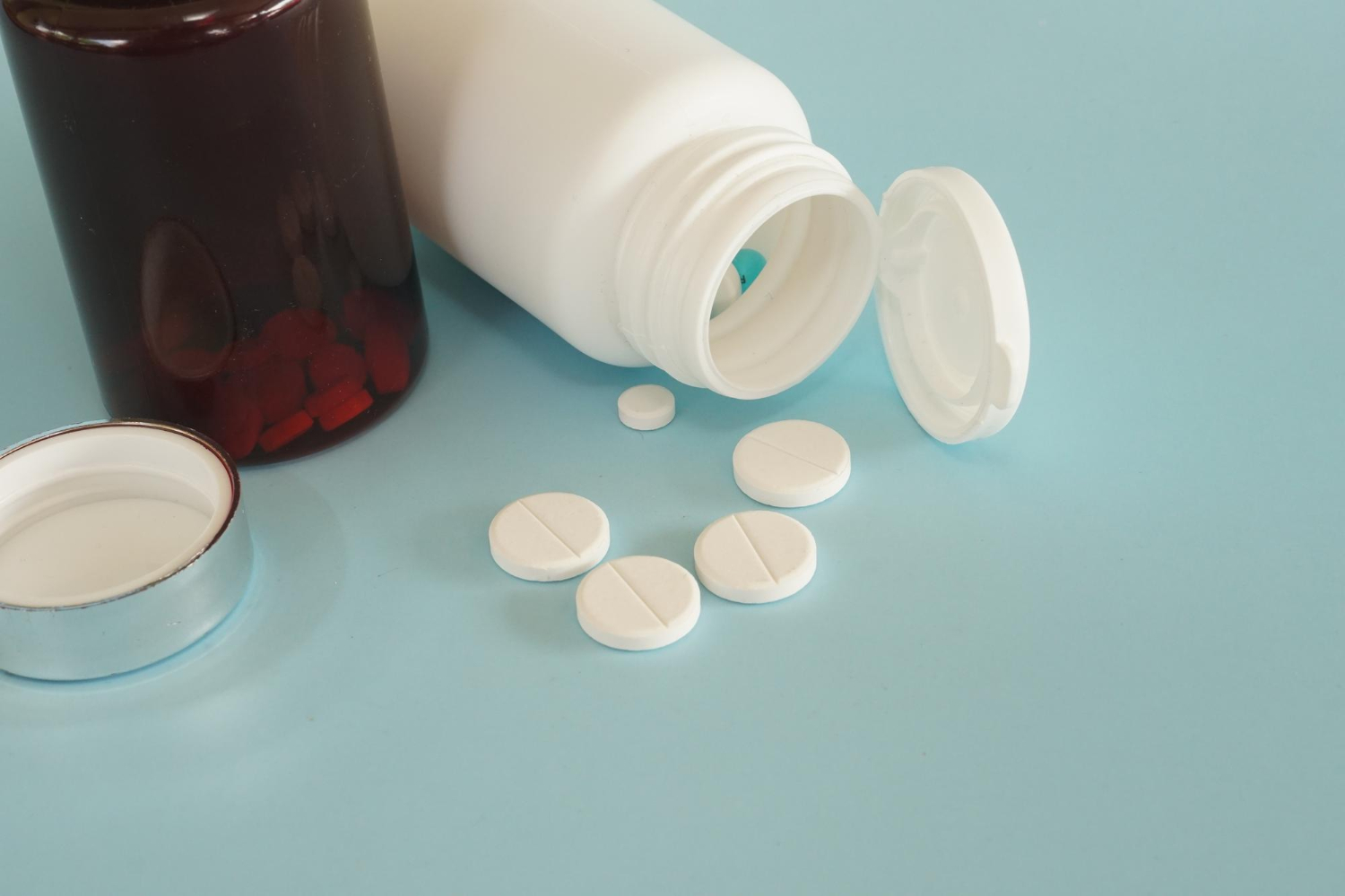 Two pill bottles on a blue surface, one open with white tablets spilled out, the other closed with red pills inside.