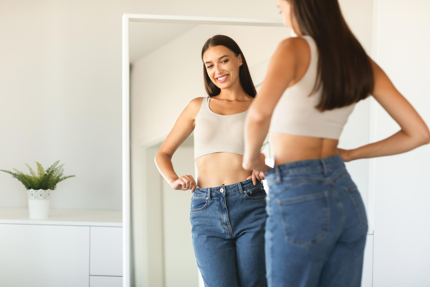 Woman smiling at her reflection in a mirror while trying on loose jeans in a bright room.